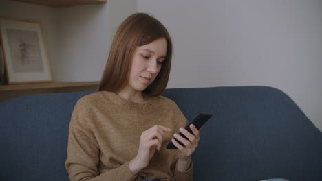 Young-woman-sitting-at-desk-at-home-office-distracted-from-work-holding-smart-phone-using-social-networking-website-chatting-with-friend-remotely-using-modern-device-and-internet-connection